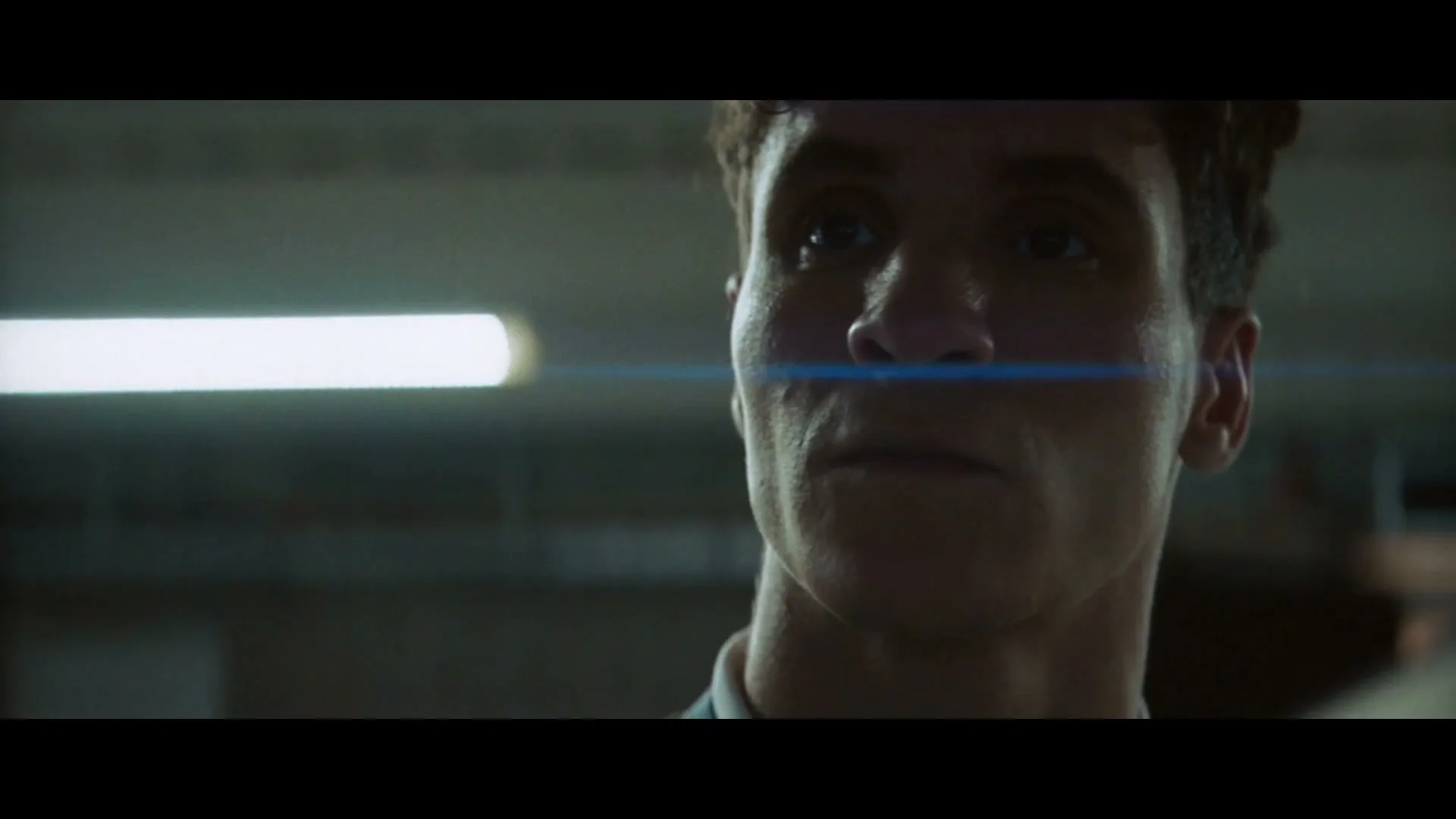 A young man with curly hair looks up, a blue lens flare crosses his face, in a dimly lit interior.