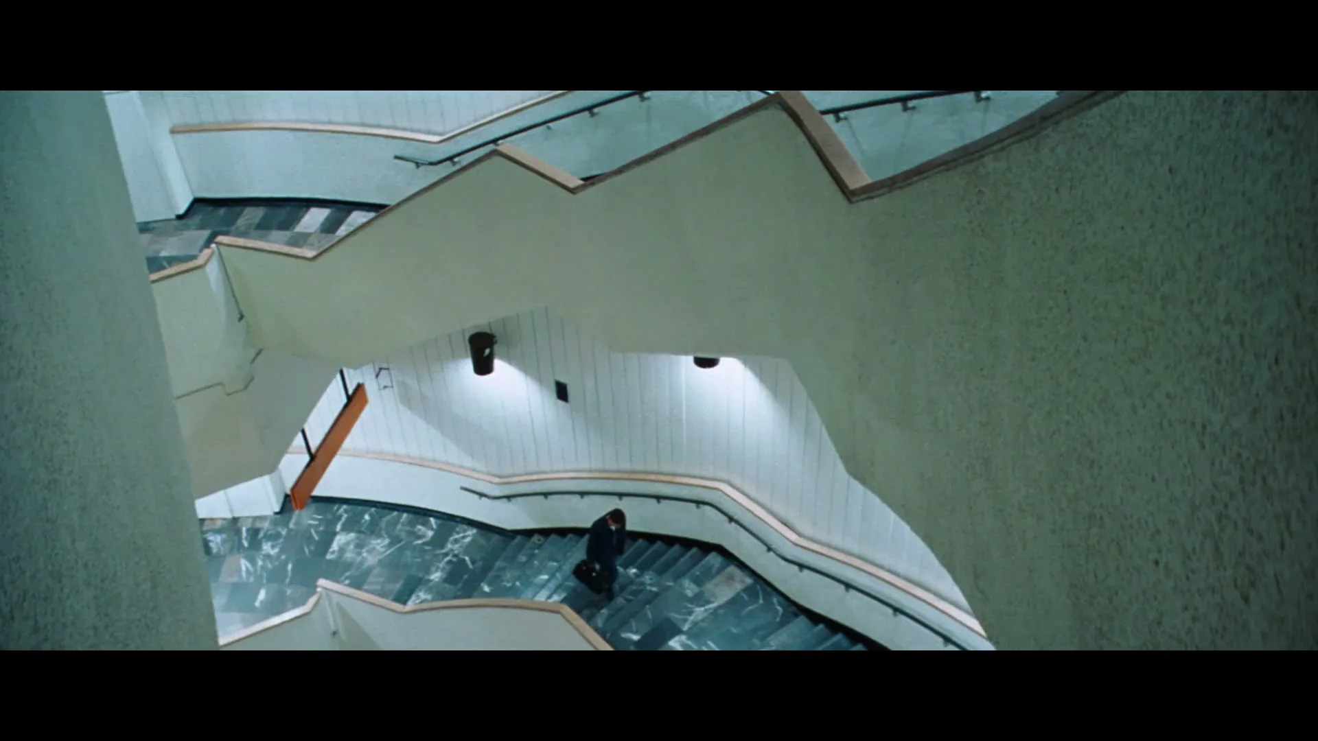 A man in a dark suit walks down a wide, modern, multi-level staircase with marble steps and white walls, seen from an overhead perspective.