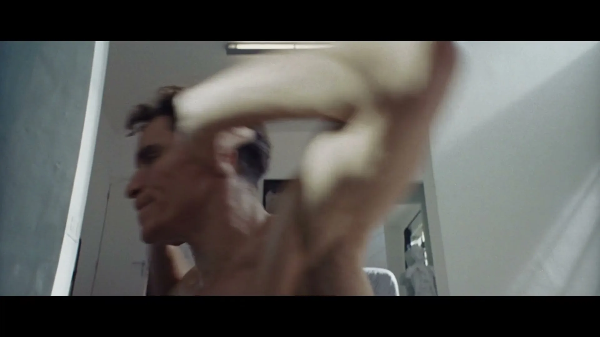 A man with short brown hair, wearing a white t-shirt, is seen from a low angle, looking up and moving his arms quickly in an interior setting.
