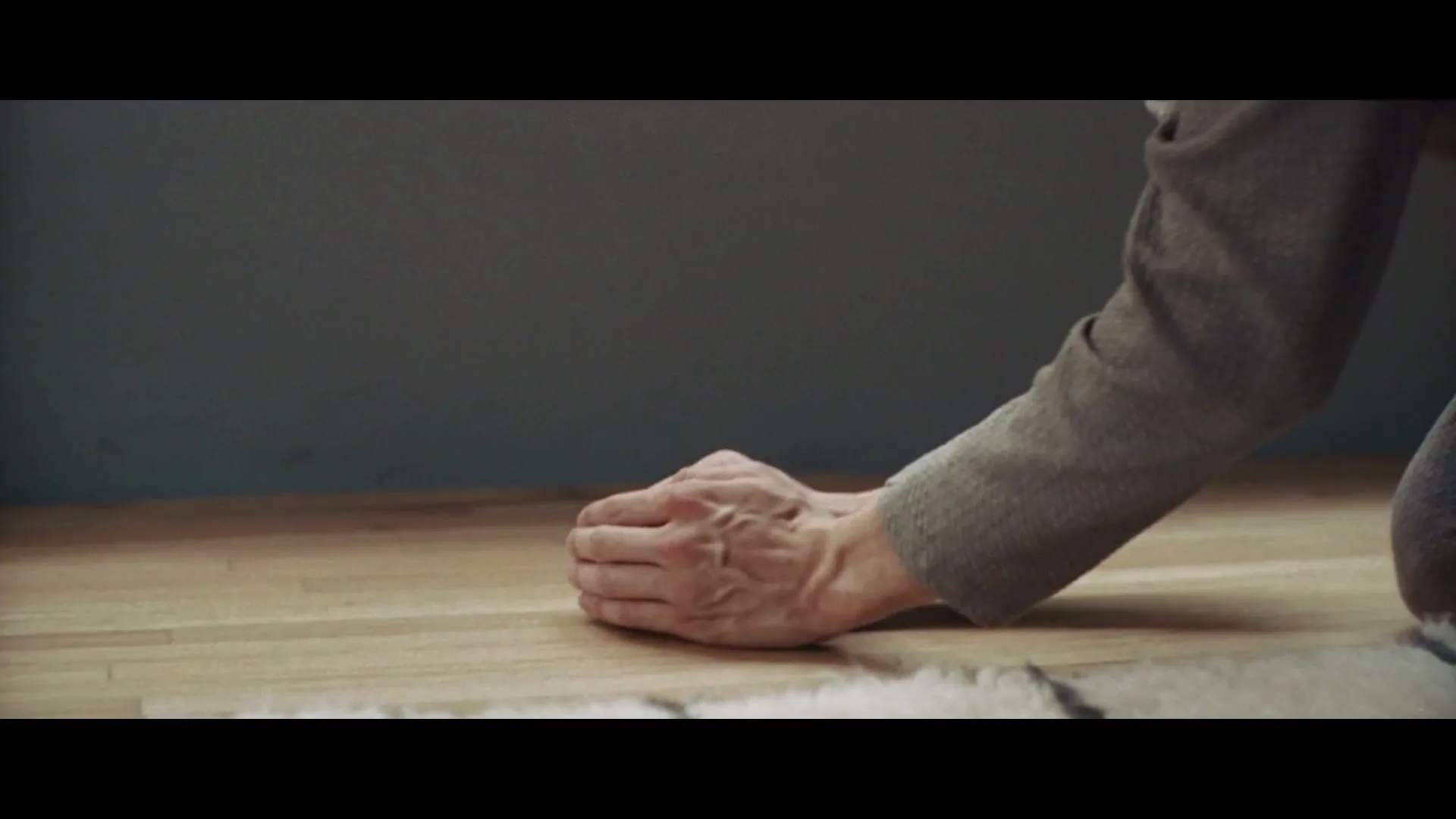 A person in a grey sweater gently cups their hands around a small bird on a light wooden floor, then closes their hands.