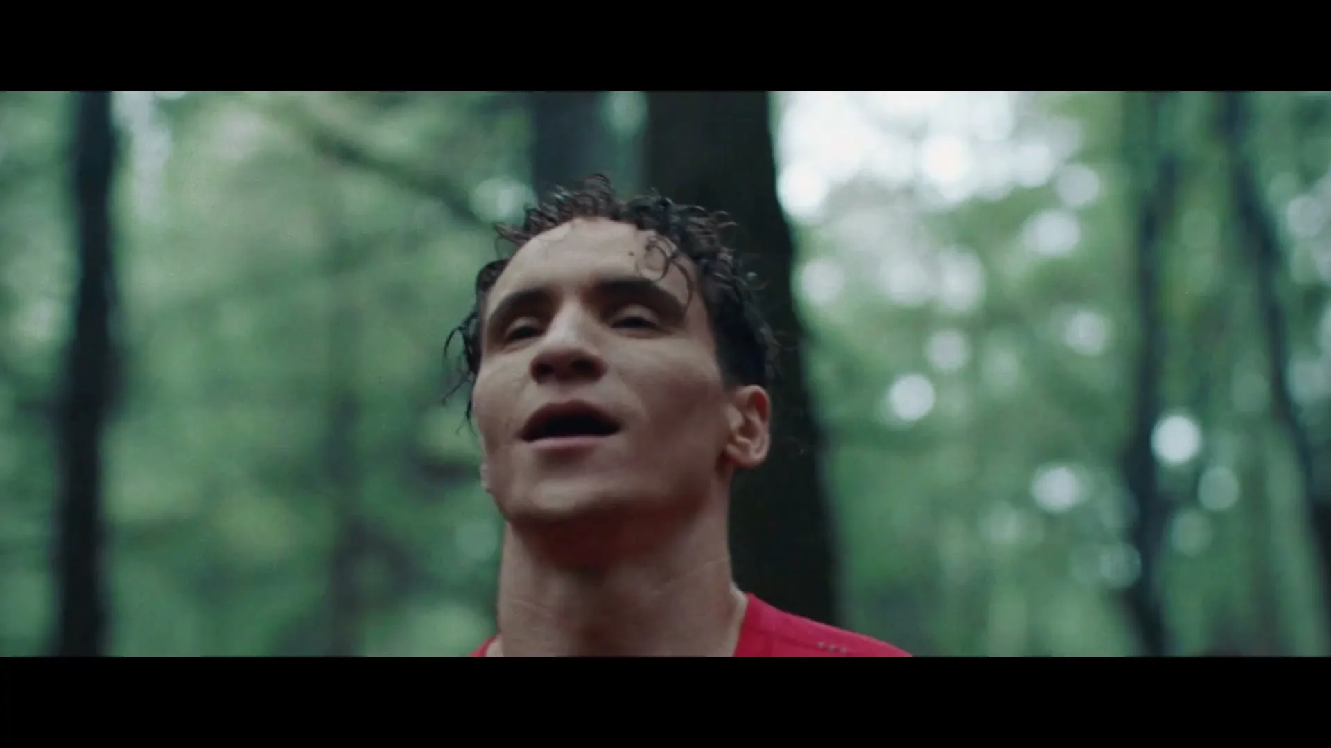 A male runner with curly hair and a red shirt looks up, sweating, as he runs through a green forest.