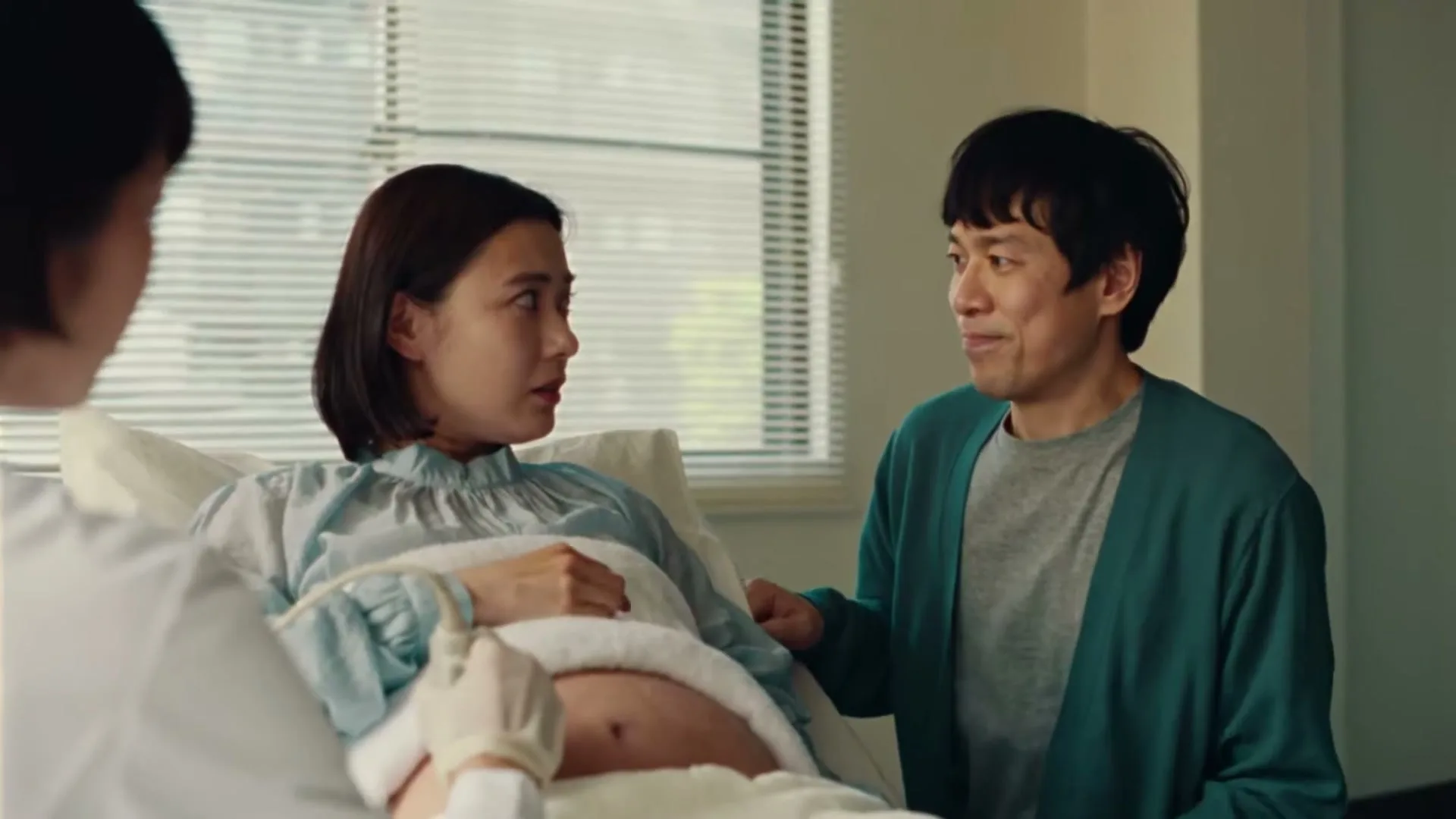 A pregnant woman lies in a hospital bed, looking concerned, while a man sits beside her, and a doctor performs an ultrasound.