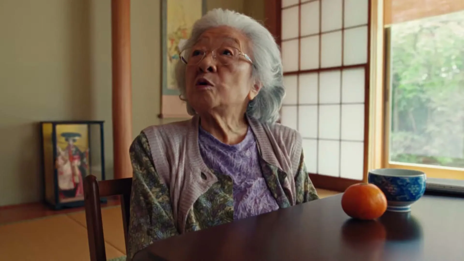 An elderly Japanese woman with white hair and glasses sits at a table, looking up with a surprised expression in a traditional Japanese room.