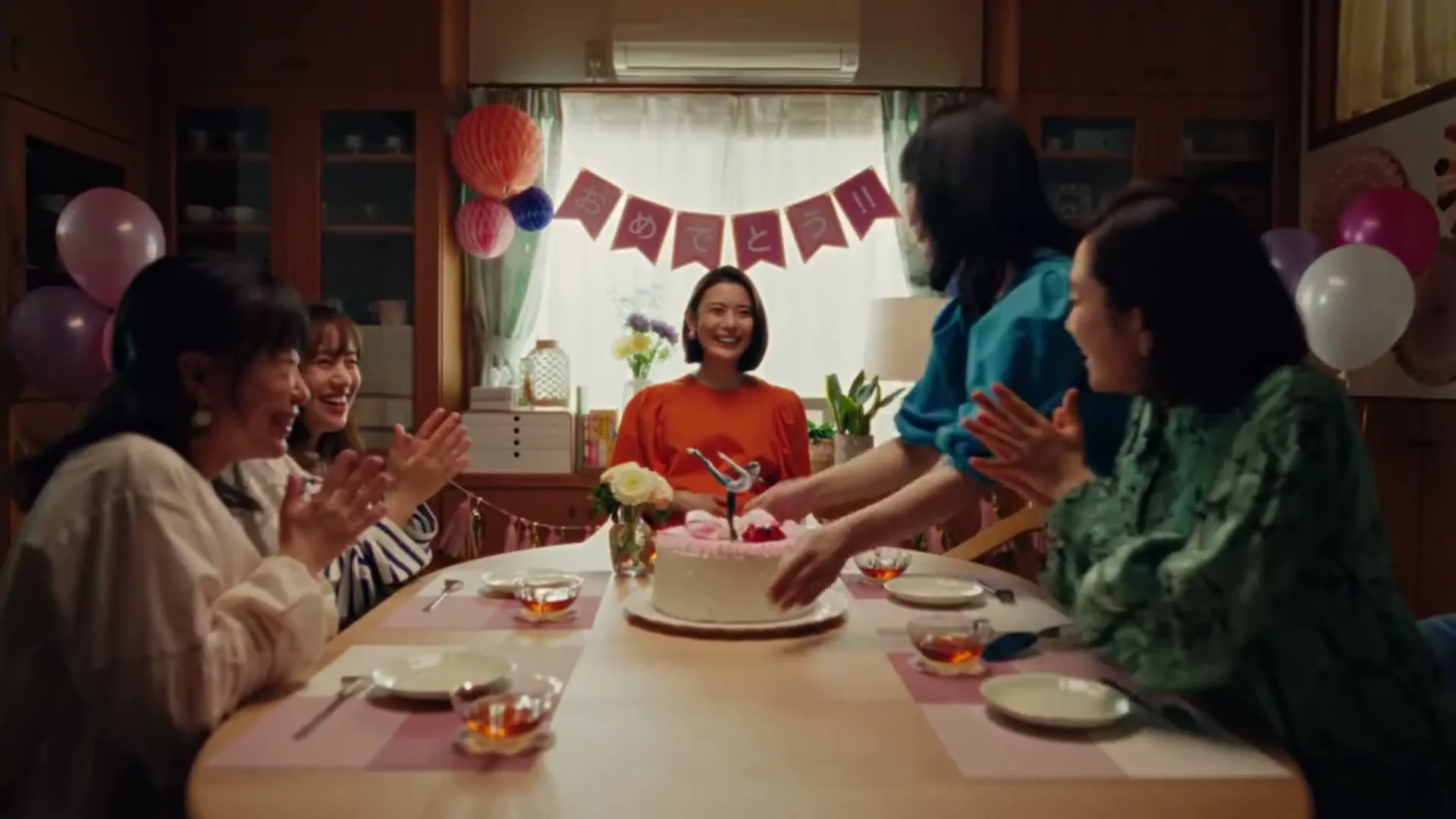 Five Japanese women celebrate a birthday, clapping around a table with a cake in a warmly lit dining room.
