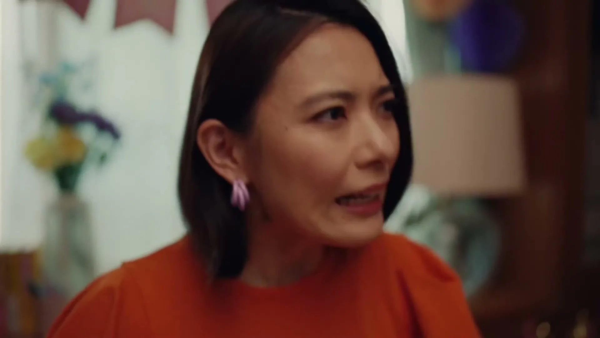 A woman with short dark hair and an orange top looks surprised and slightly annoyed in a decorated room.
