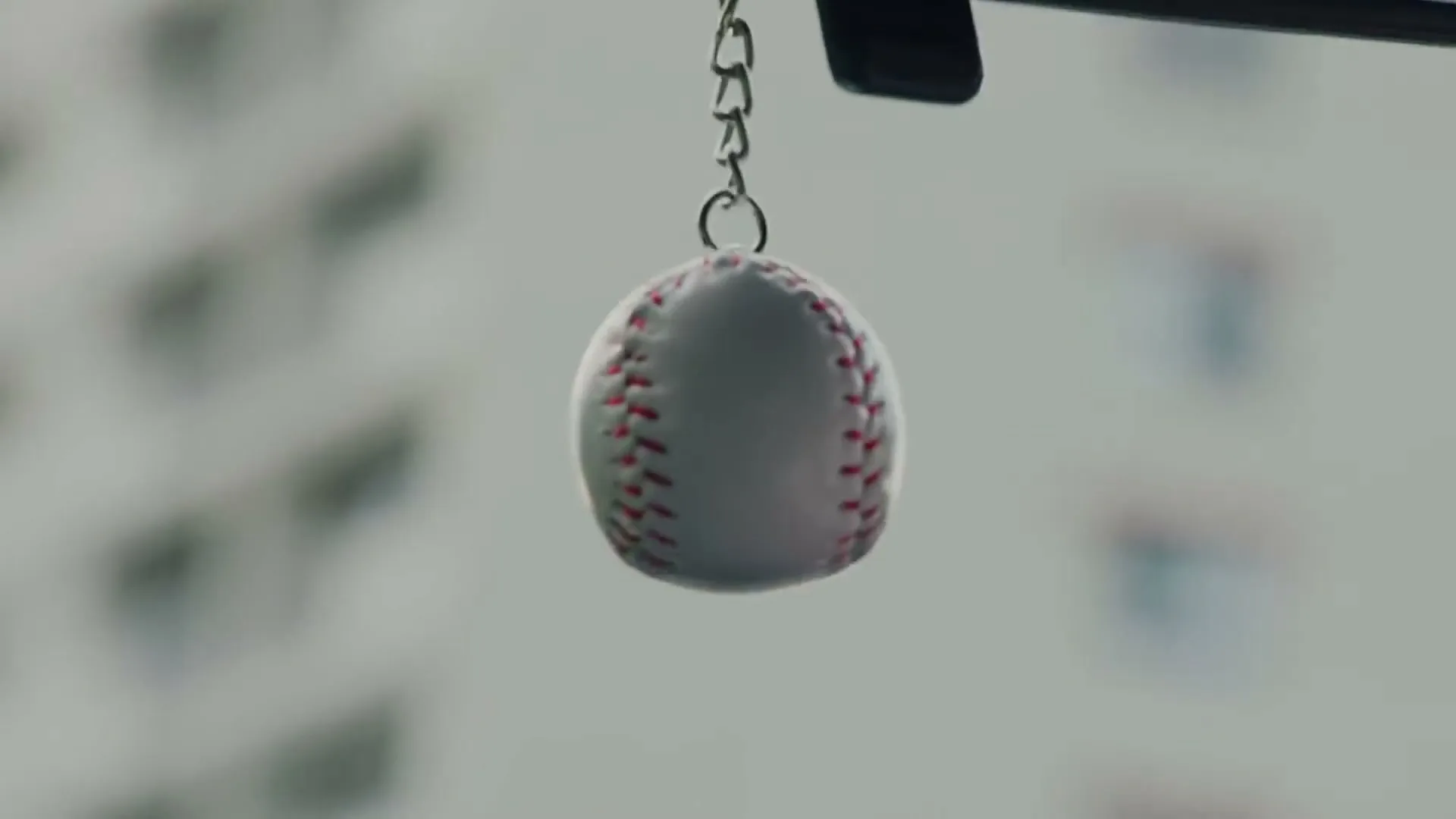 A small baseball keychain hangs from a car's rearview mirror, with a blurred city building in the background.