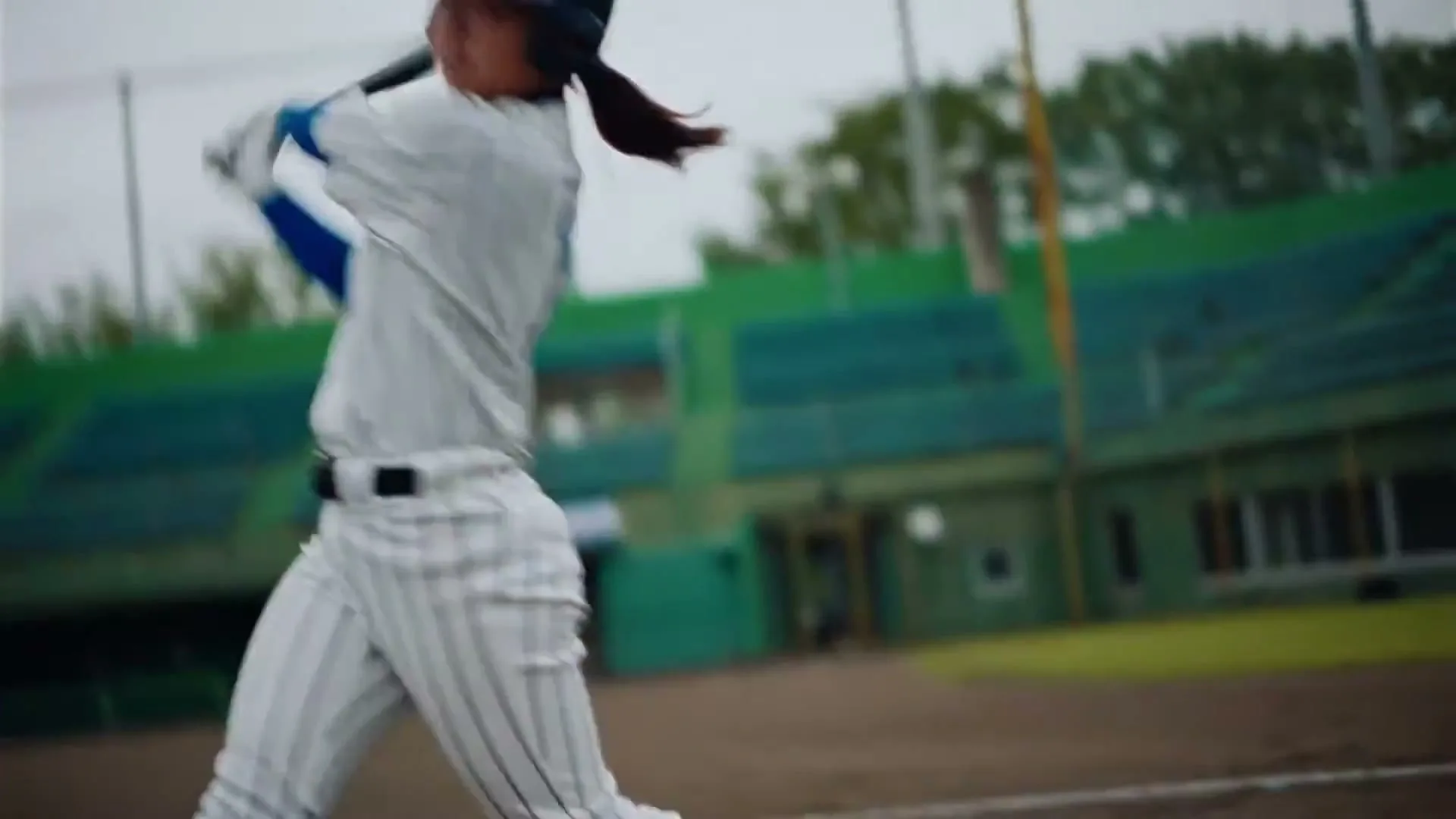 A female baseball player in a white and blue uniform swings a bat with motion blur on a baseball field during the day.