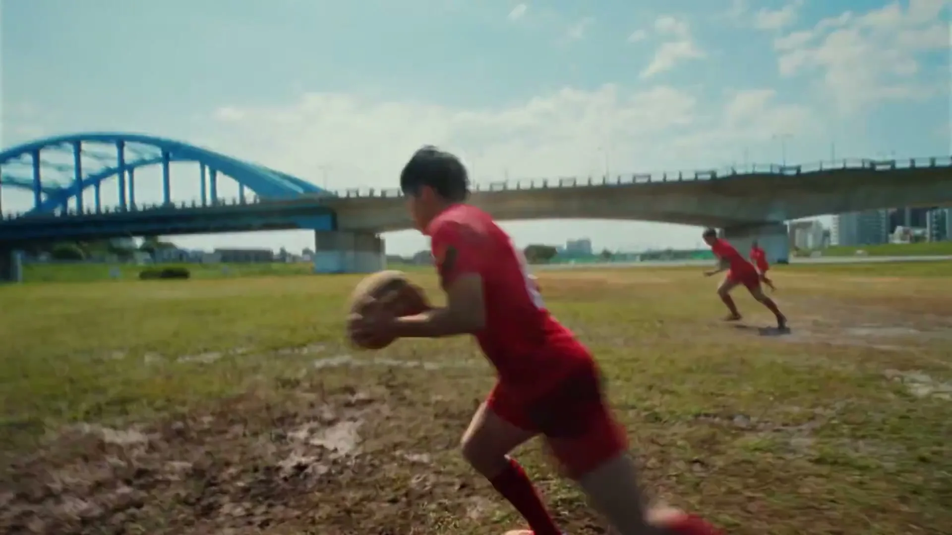 A young female rugby player in a red uniform runs with the ball across a muddy field under a blue sky, with a bridge in the background.