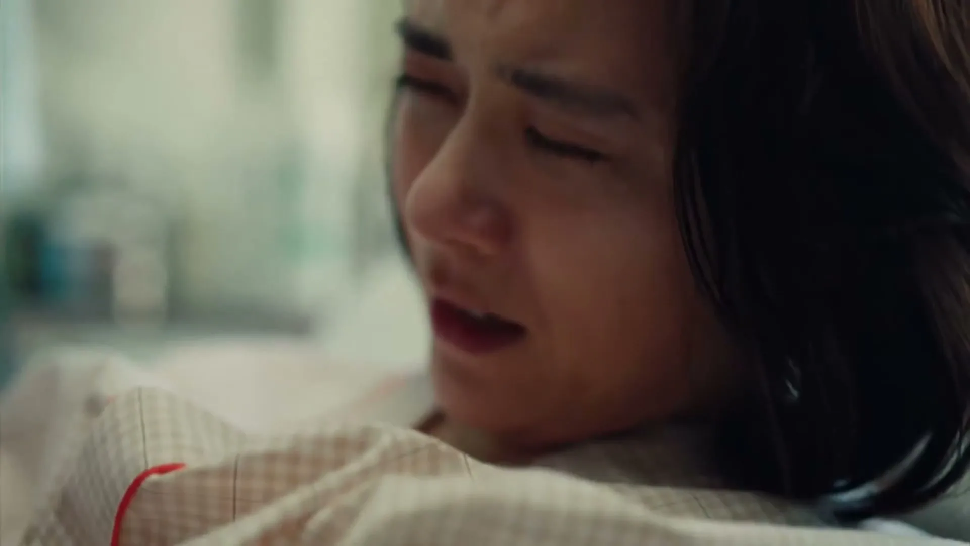 A woman with dark hair and a white shirt is seen in a medium closeup, looking distressed and in pain, possibly in a hospital bed.