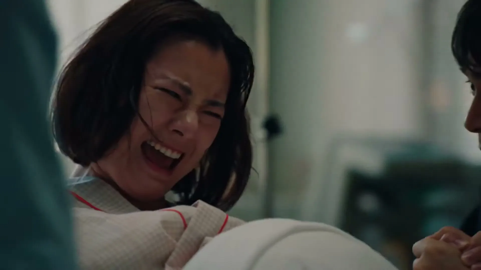 A woman in a hospital gown screams in pain while giving birth, supported by another person in a hospital room.