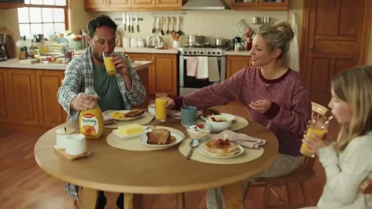 A family of three, a man, woman, and child, are having breakfast in their kitchen, pouring and drinking orange juice.