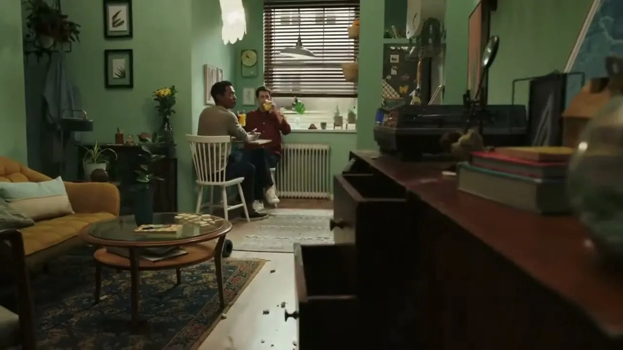 Two men are sitting at a small table in a mint green apartment, one drinking from a glass, the other looking at him.