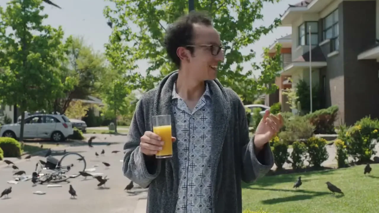 A man in a bathrobe and glasses stands on a suburban street, holding orange juice, surrounded by crows and a fallen bicycle.