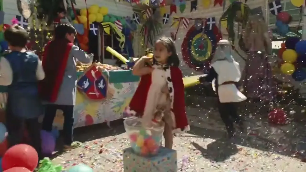 A young girl in a red cape stands in a confetti-filled backyard, surrounded by other children in costumes at a lively party.