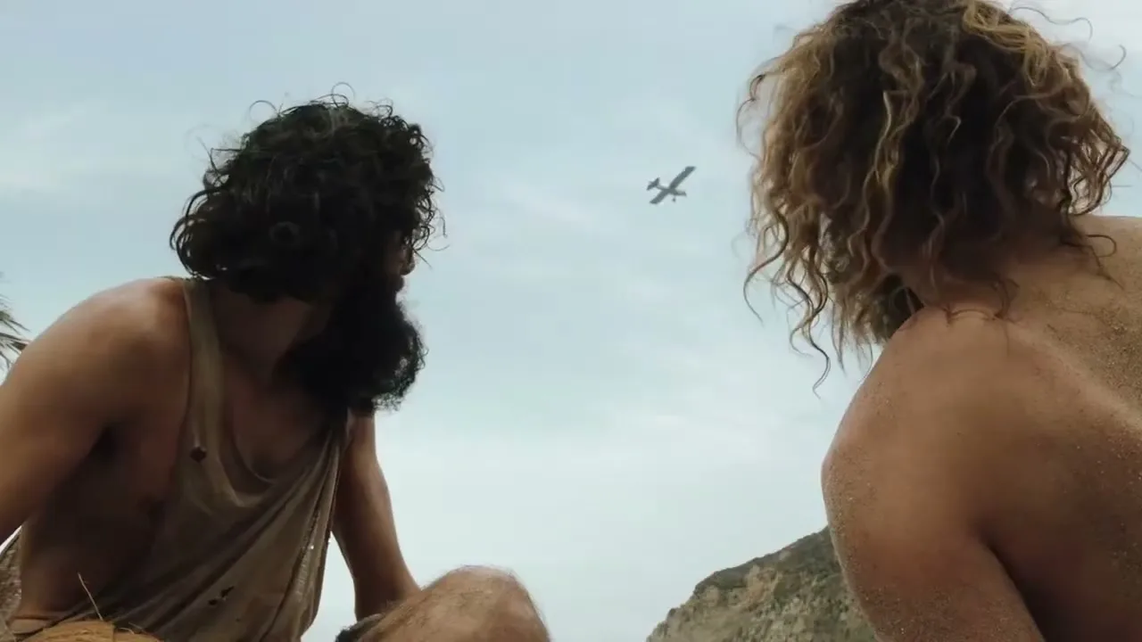 Two men with long, curly hair, covered in sand, look up at a small plane flying across a bright, overcast sky on a beach.