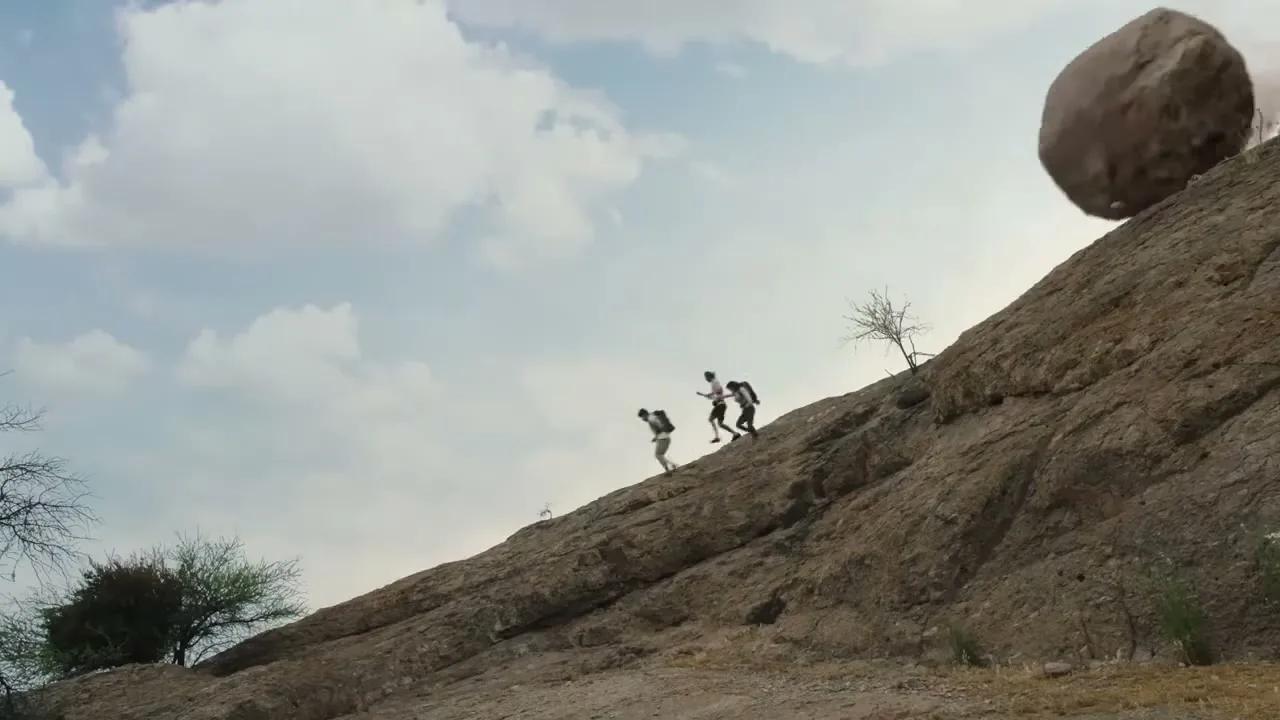Three people in casual clothes run uphill on a rocky slope as a large boulder rolls down towards them under a cloudy sky.
