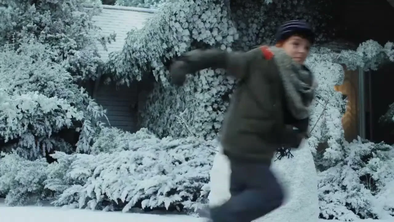 A young boy in winter clothes throws a snowball at a snowman in a snowy suburban yard.