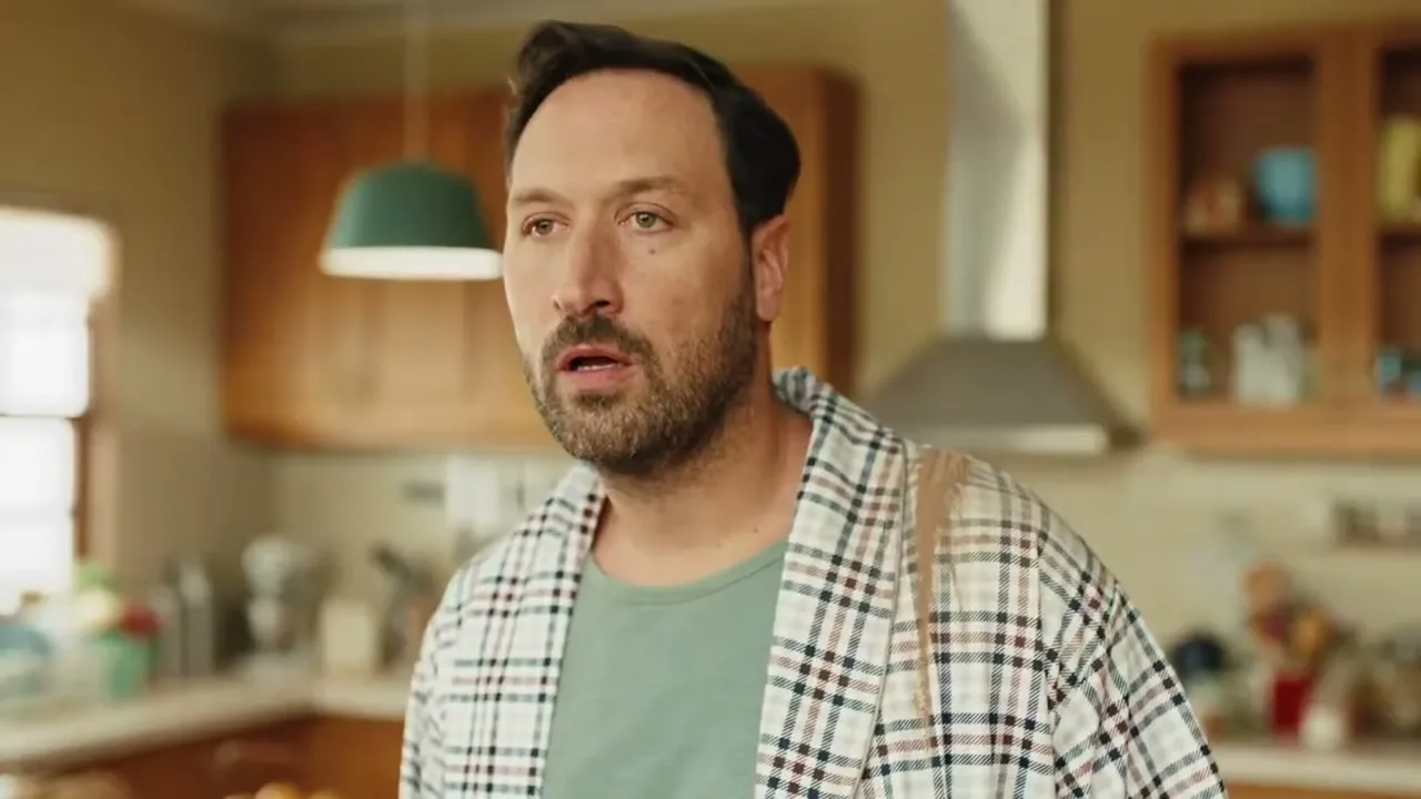 A man in his 30s, wearing a plaid robe, looks surprised in a brightly lit kitchen during the day.