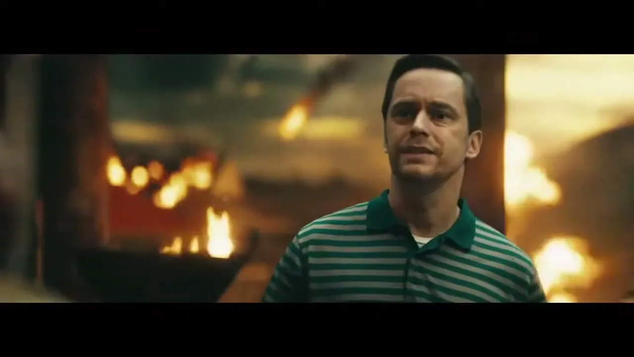 A man in a striped polo shirt looks around with a concerned expression amidst a fiery, chaotic background, possibly a disaster scene.