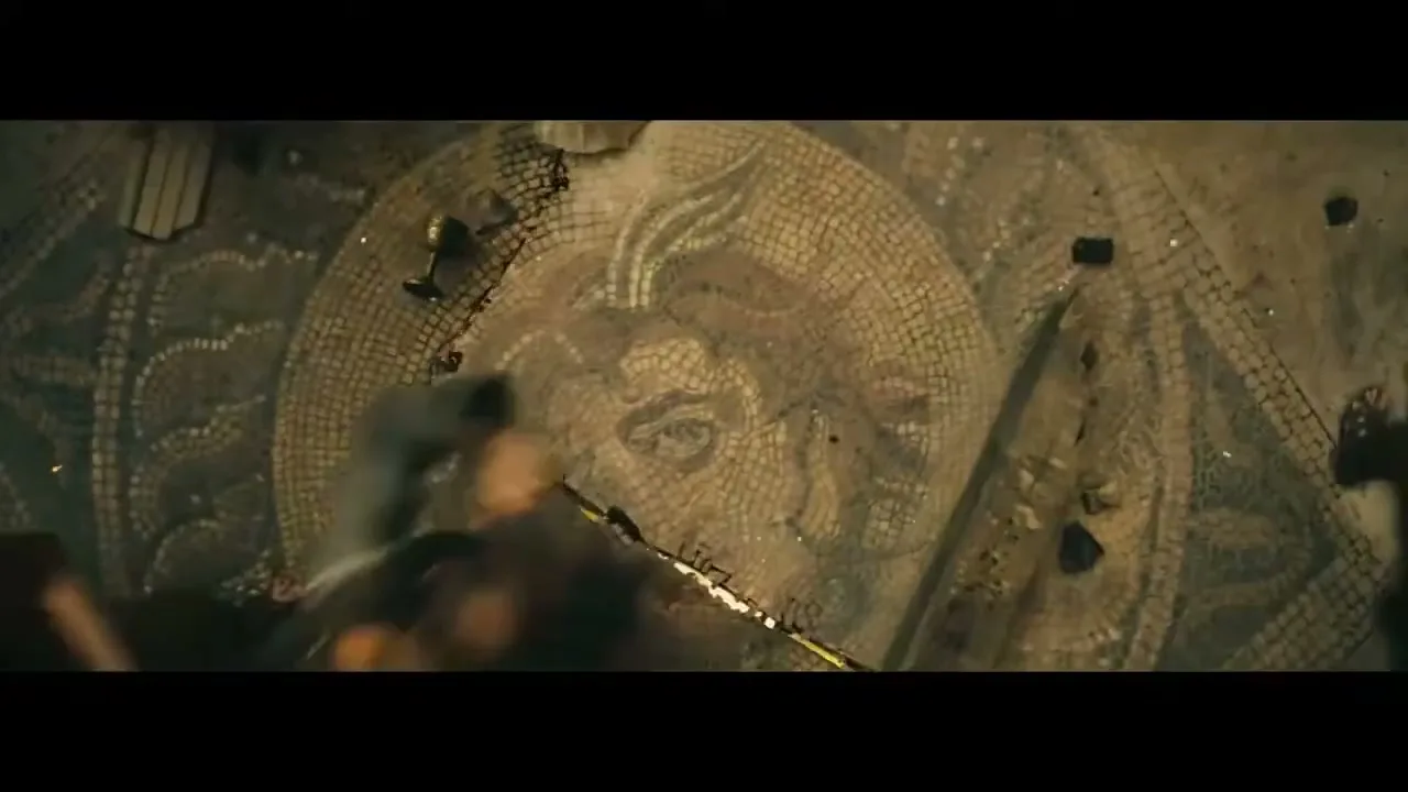 A high-angle shot shows a detailed, ancient mosaic of a face on the floor, as a person in a dark tunic walks across it, kicking up debris.