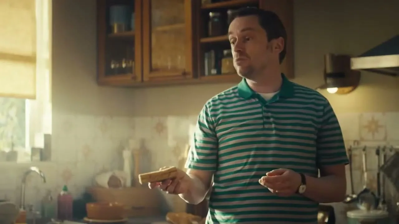 A man in a green striped shirt holds a piece of toast and looks off-camera, standing in a sunlit kitchen.