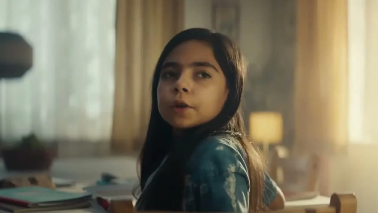 A young girl with long dark hair, wearing a blue tie-dye shirt, turns her head to look at someone off-screen in a sunlit room.