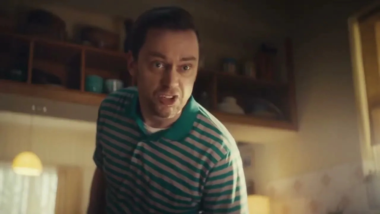 A man in his 30s, wearing a green and white striped polo shirt, looks around a kitchen with a worried expression, as if searching for something.