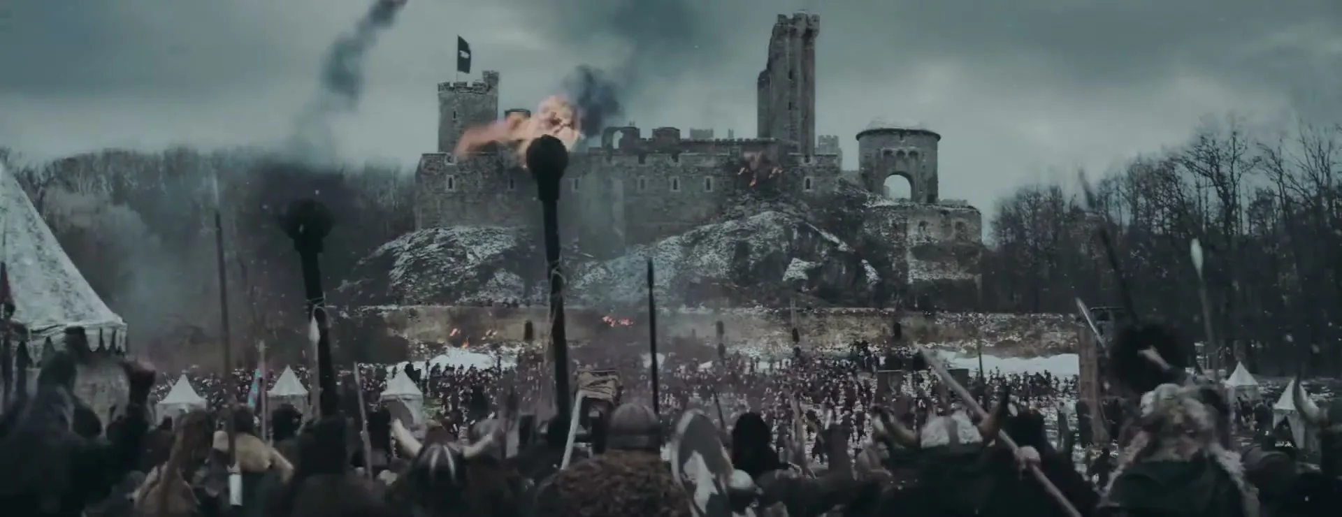 A wide shot shows a medieval castle under siege, with a large army of warriors in the foreground and smoke rising from the castle.