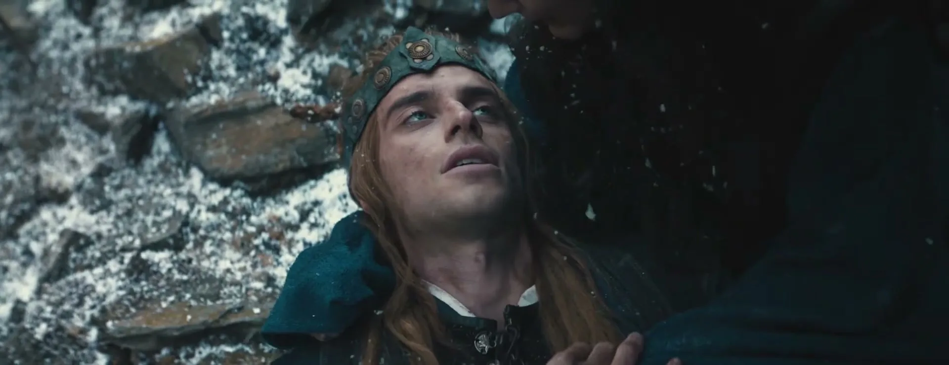 A young man with long blonde hair and a crown lies on snowy ground, looking up at someone out of frame, as snow falls around him.