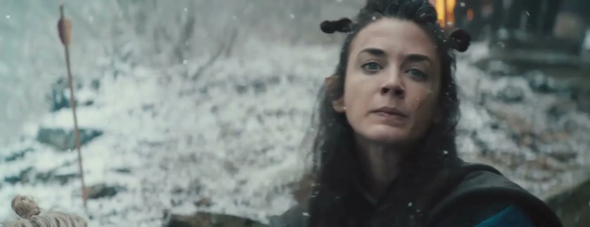 A woman with dark hair and elf ears, wearing dark clothing, looks up in pain, then directly at the camera, in a snowy, rocky landscape.