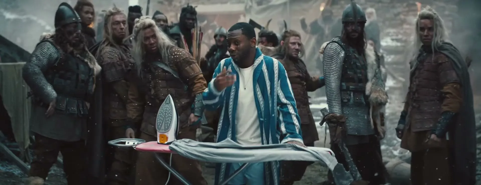 A man in a blue and white striped robe irons a shirt on an ironing board, surrounded by a crowd of Vikings in a snowy, war-torn village.