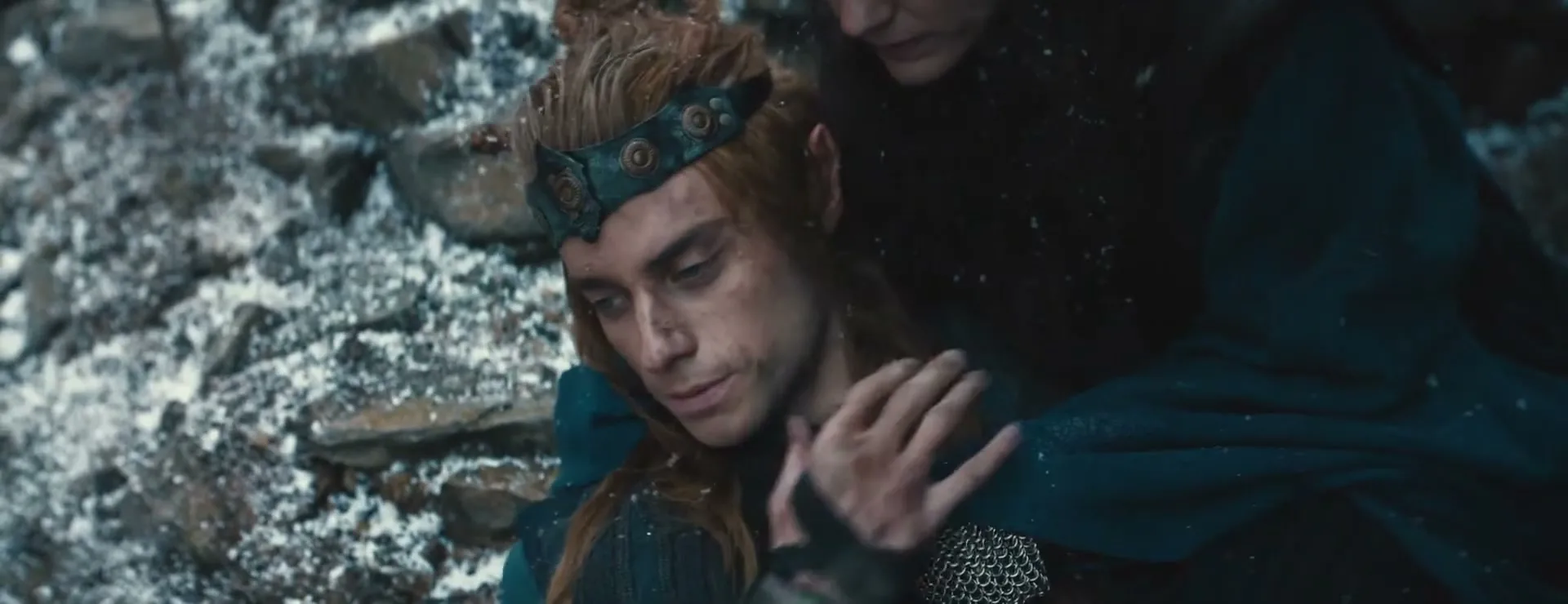 A male elf with long red hair and a crown is held by another person in a snowy, rocky landscape as snow falls.