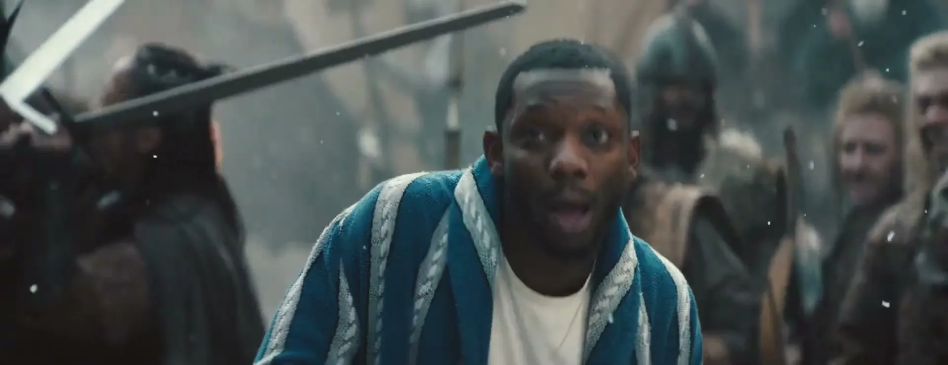 A Black man in a blue and white striped robe looks shocked amidst a chaotic medieval battle scene with warriors clashing swords.