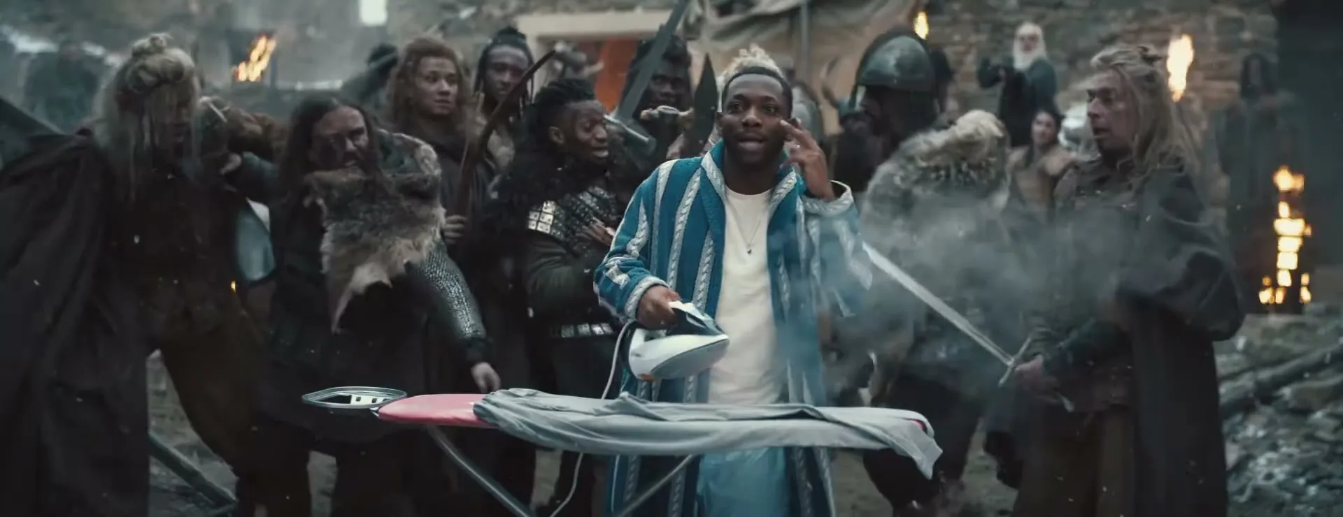 A Black man in a blue and white striped robe irons a grey shirt on an ironing board, surrounded by a crowd of Vikings in a ruined village.