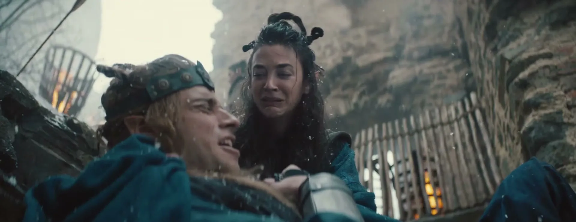 A woman with dark hair and a man with long blonde hair and elf ears are in a fantasy setting, with the woman holding the man as snow falls around them.