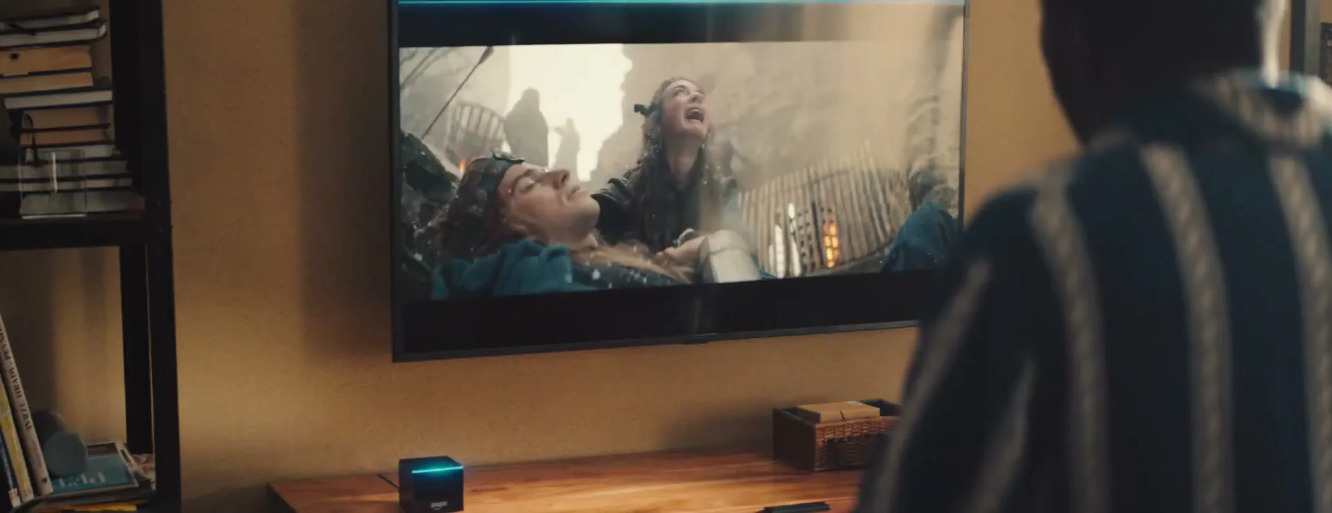 A man watches a fantasy movie on a TV, with a smart speaker on the table below, in a living room.