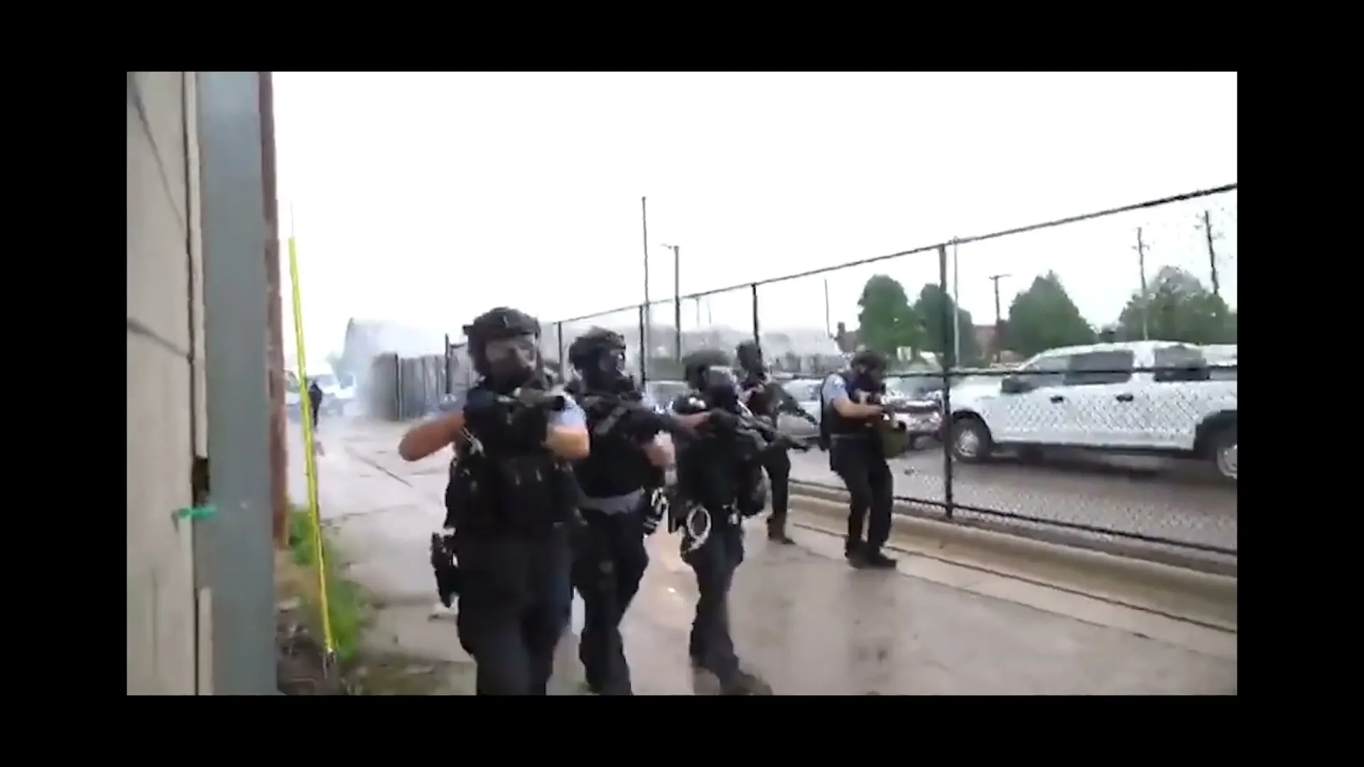 A group of police officers in tactical gear and gas masks hold rifles and advance down a wet alleyway next to a chain-link fence on a cloudy day.