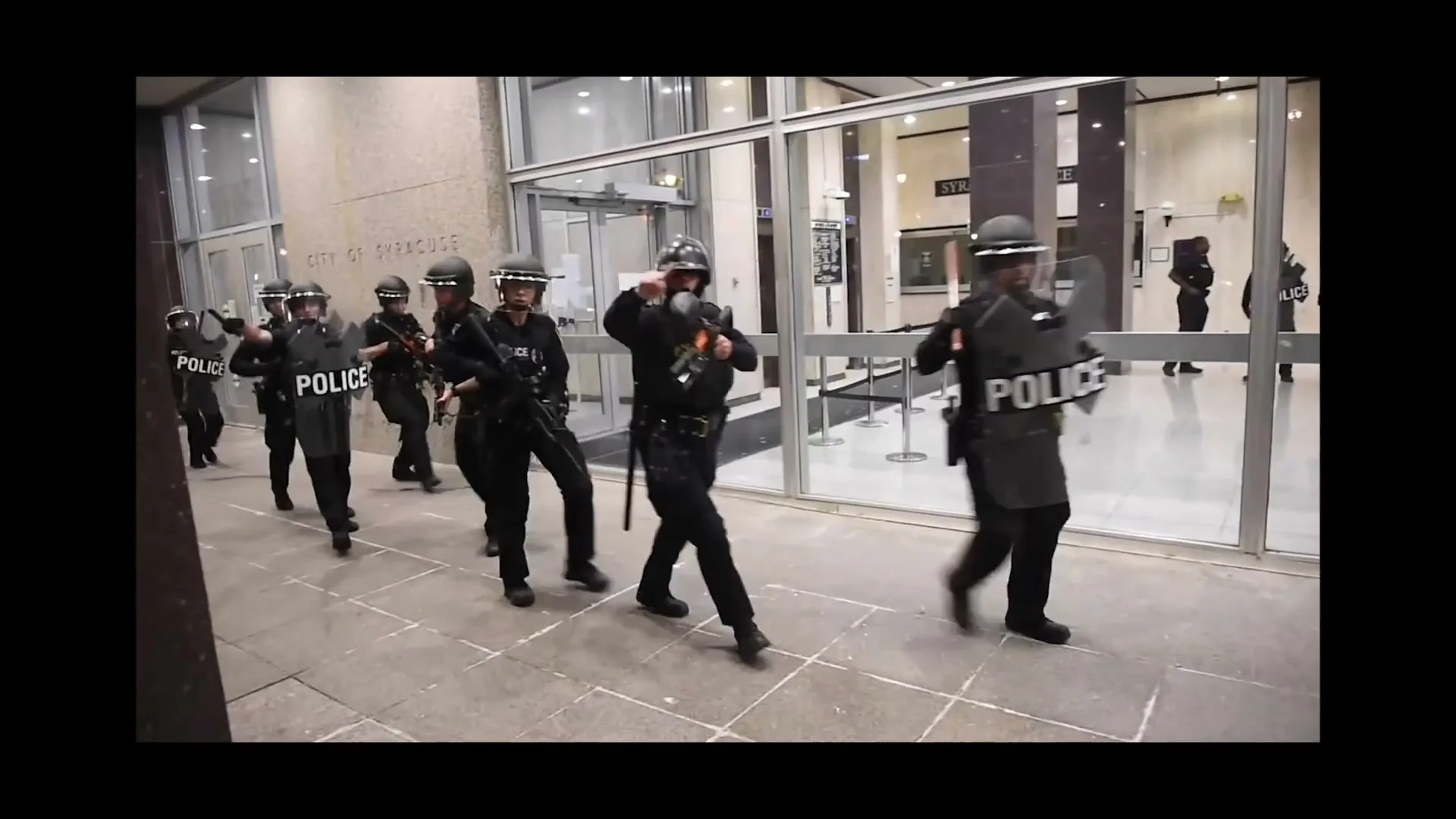 A group of police officers in riot gear run forward, some holding shields, in front of a modern building with large glass windows.