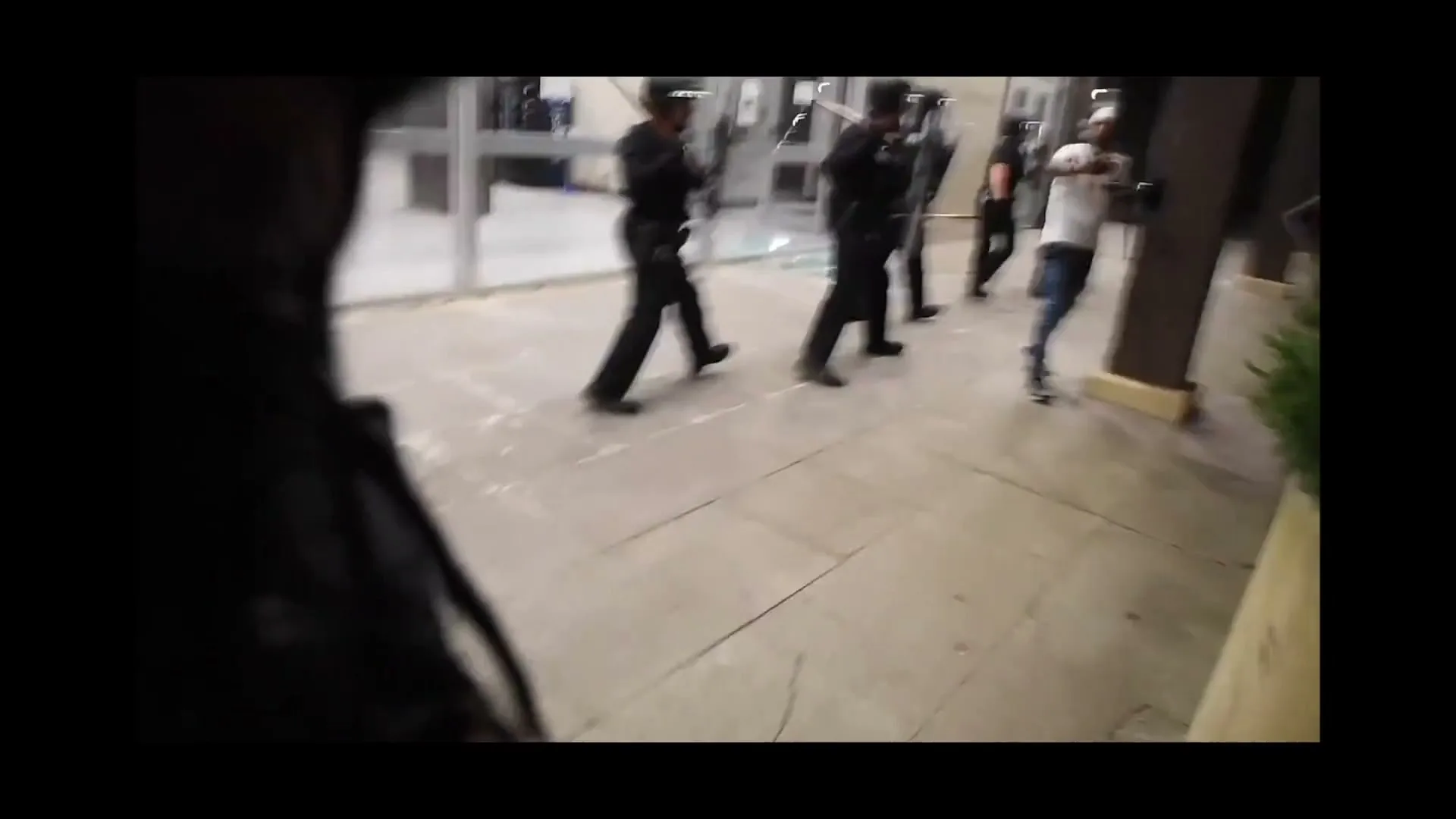 A group of police officers in riot gear with shields and batons run forward, while a civilian runs away from them in an interior space.
