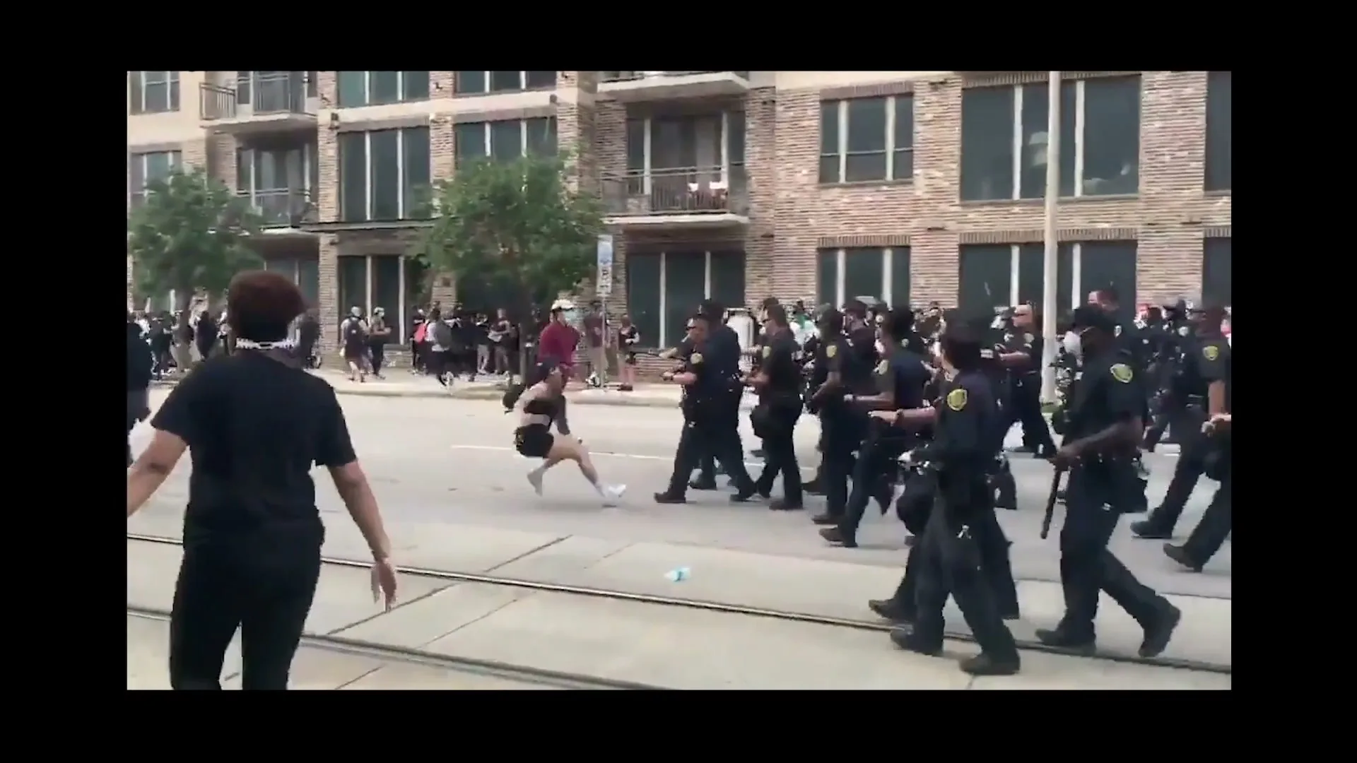 A woman in a black top and shorts is pushed to the ground by police officers during a protest on a city street.