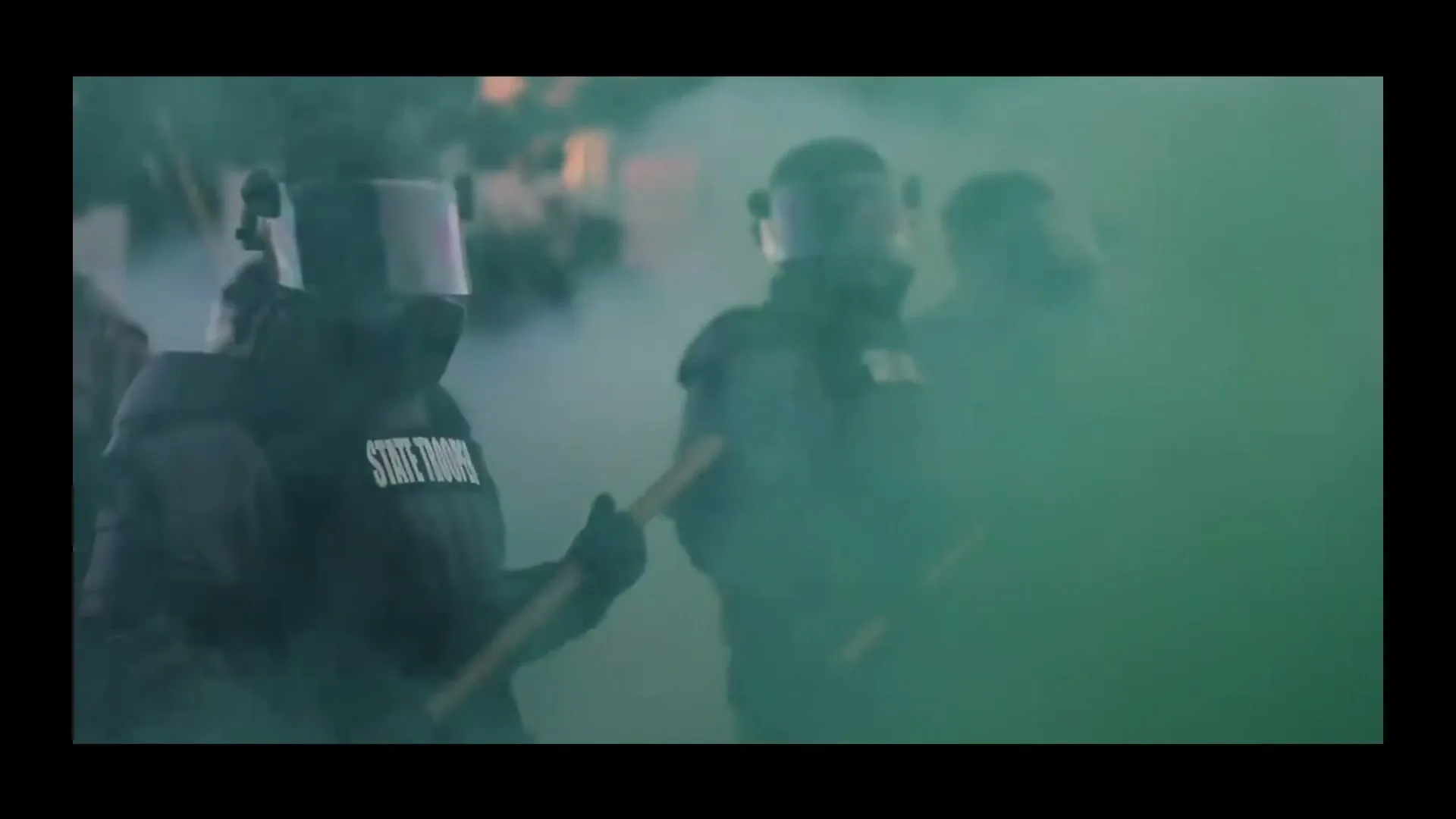 Three state troopers in riot gear and helmets stand in a cloud of green smoke, holding batons during a protest.