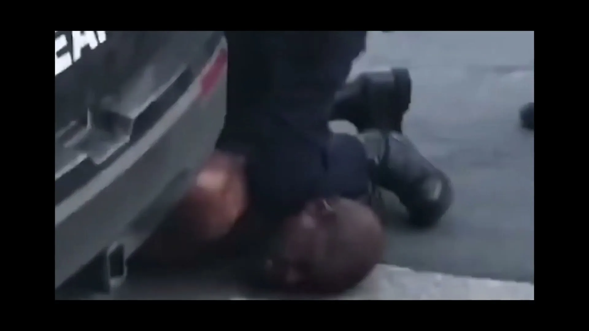 A Black man is pinned to the ground by a police officer's knee, next to a car on a street.