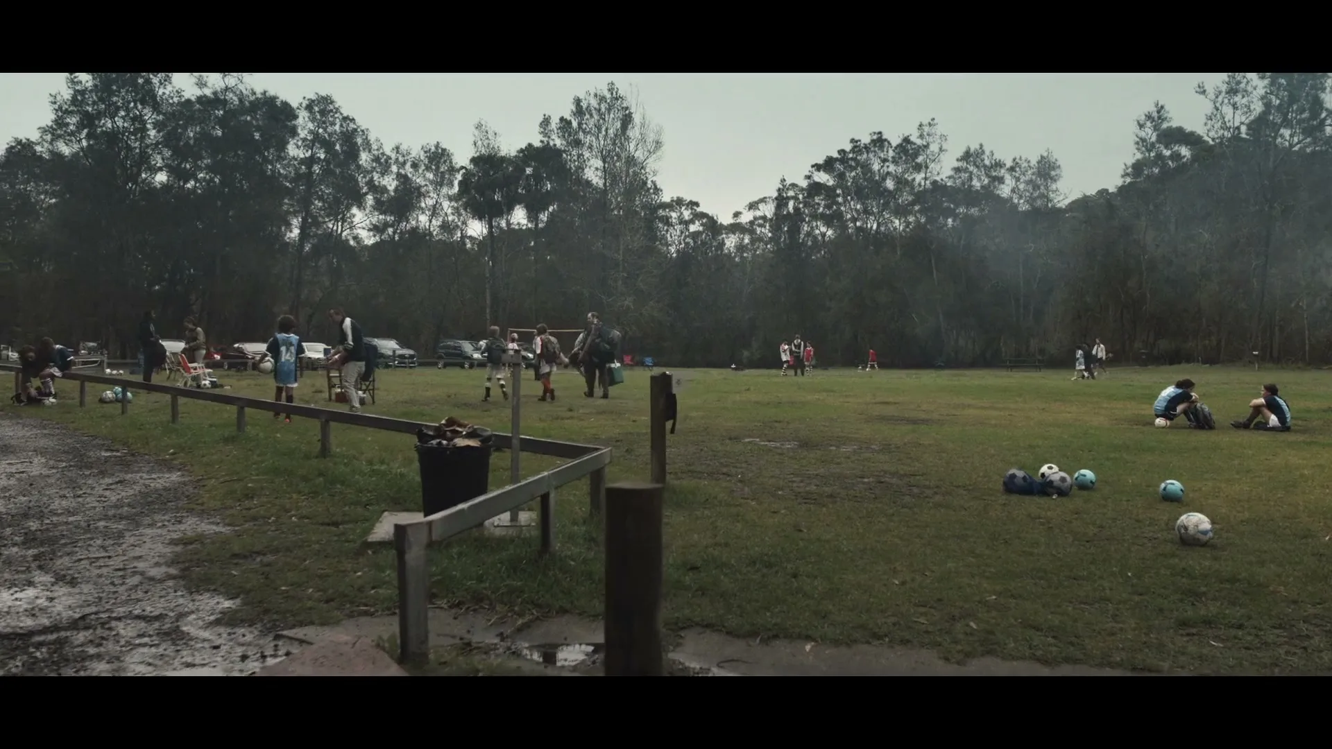 A wide shot of a muddy soccer field with children and adults scattered around, some playing, some resting, under an overcast sky.