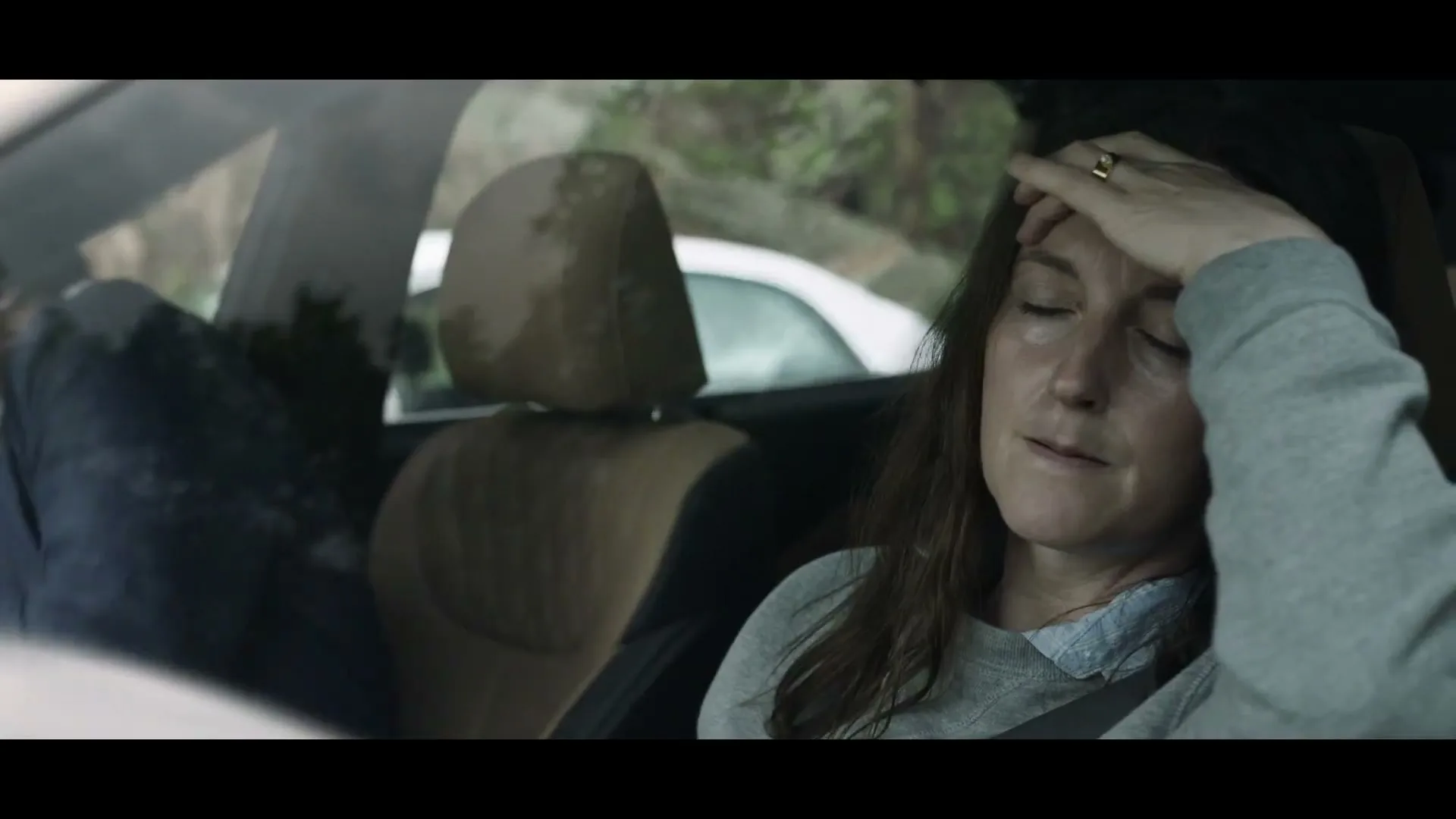 A woman with long brown hair, wearing a grey sweatshirt, sits in the passenger seat of a car, looking stressed with her hand on her forehead.