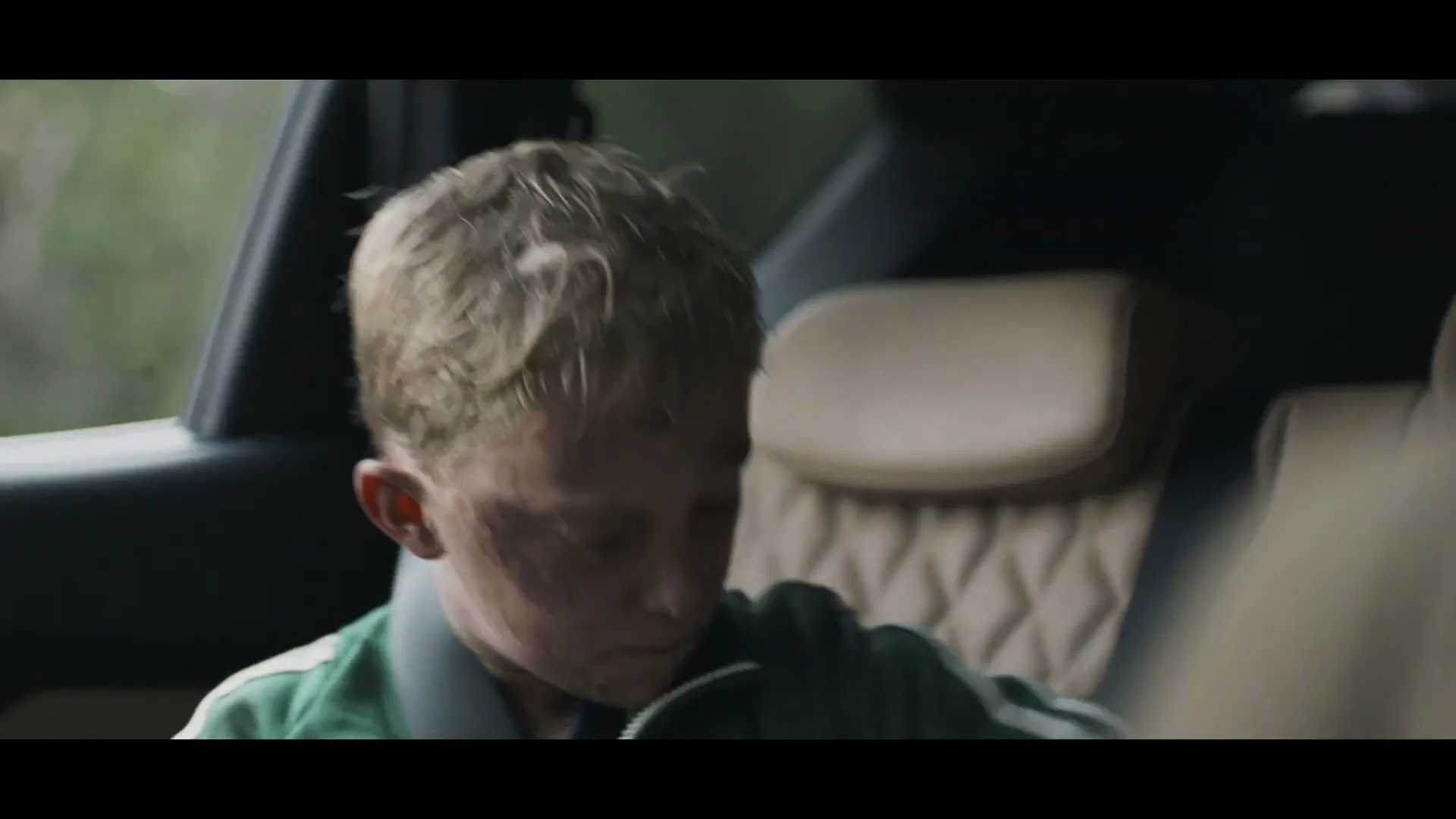 A young boy with dirty face and blonde hair, wearing a green tracksuit, looks around in the back seat of a car.