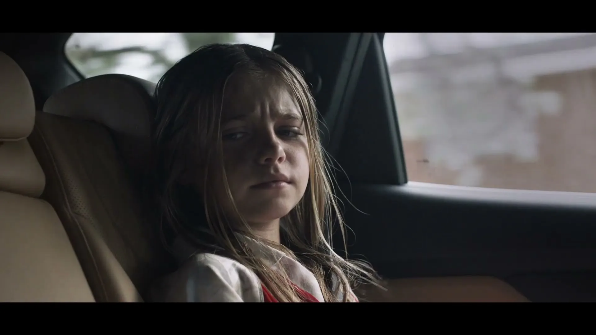 A young girl with wet hair sits in the back seat of a car, looking sad and worried.