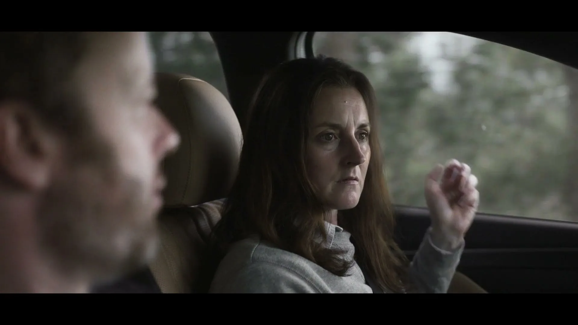 A woman with brown hair in a grey top looks out the car window, then turns to look at the man driving, in a car on a cloudy day.
