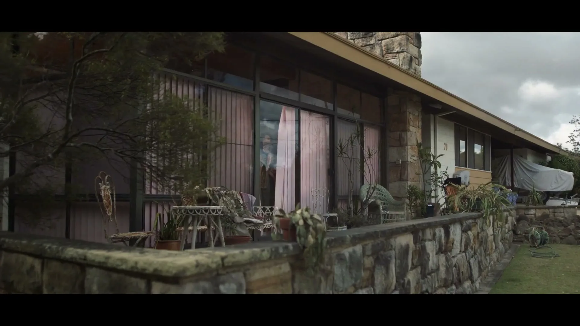 A woman in a green dress stands in the doorway of a modern house with a stone wall and pink curtains, looking out.