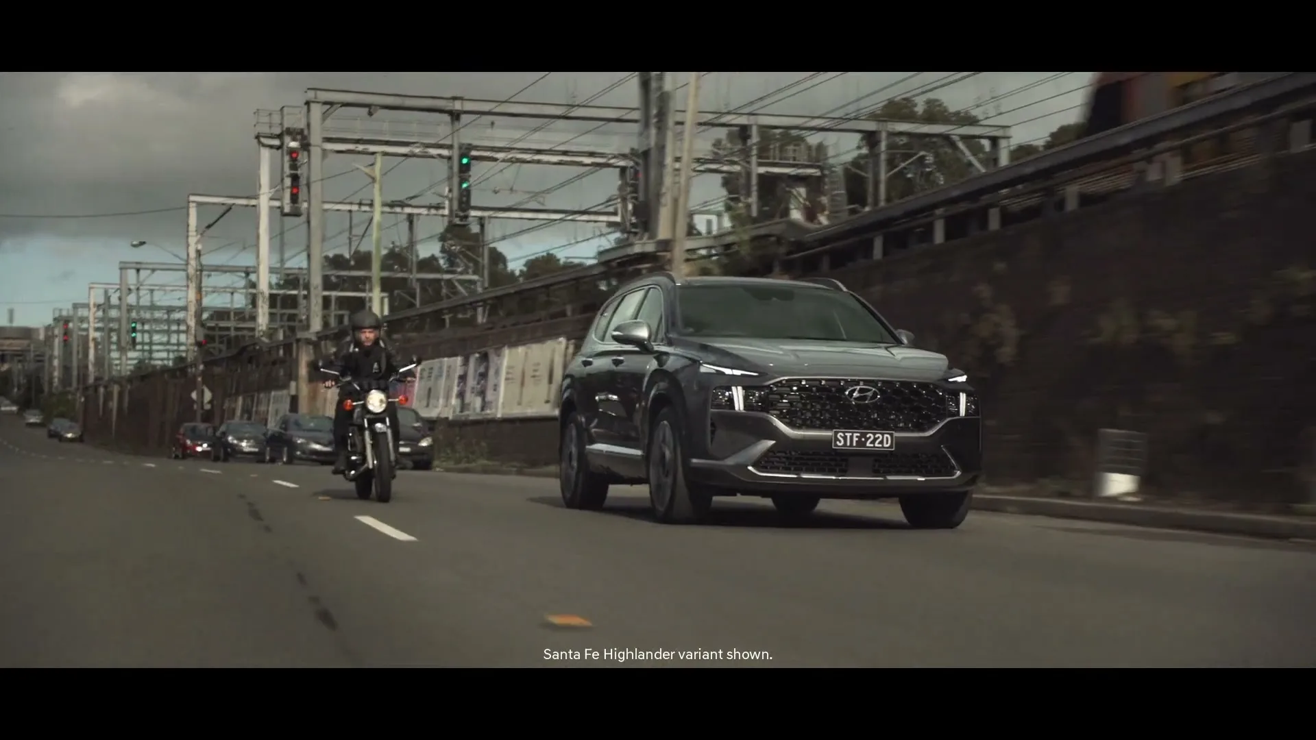 A dark grey Hyundai Santa Fe SUV drives on a multi-lane road, with a person on a motorcycle to its left, and a train passing on an elevated track in the background.