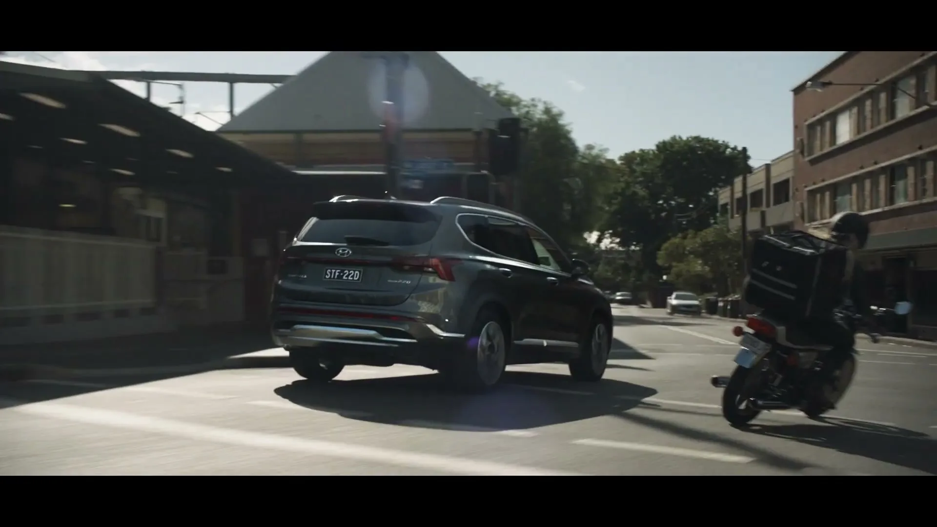 A dark grey Hyundai Santa Fe SUV drives on a city street, followed by a person on a motorcycle with a delivery box.